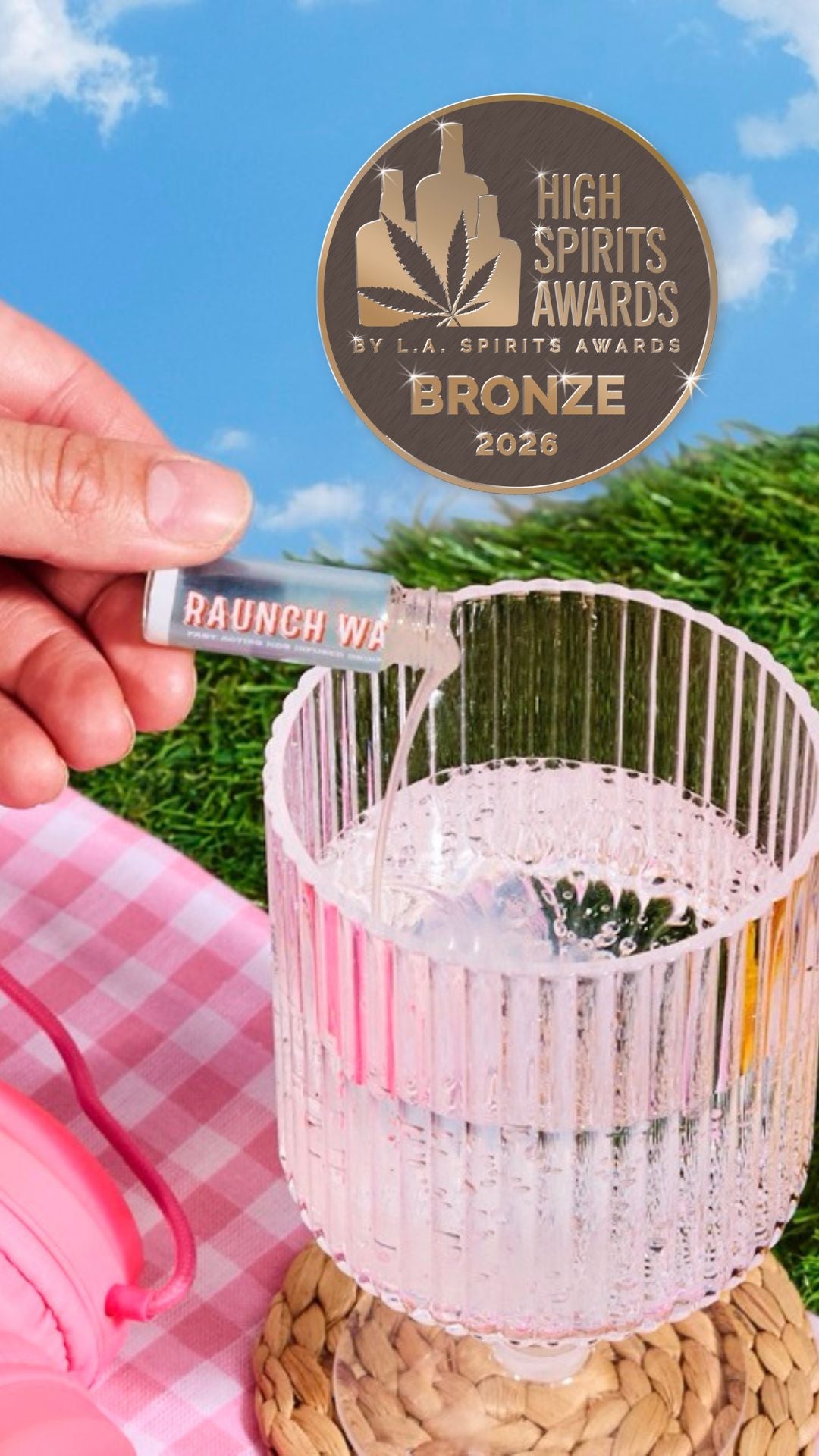 A hand pours a clear liquid from a small clear bottle labeled 'RAUNCH WATER' into a ribbed clear glass on a woven coaster, with a pink gingham cloth, grass, blue sky, and a bronze 'High Spirits Awards 2026' badge in the background.