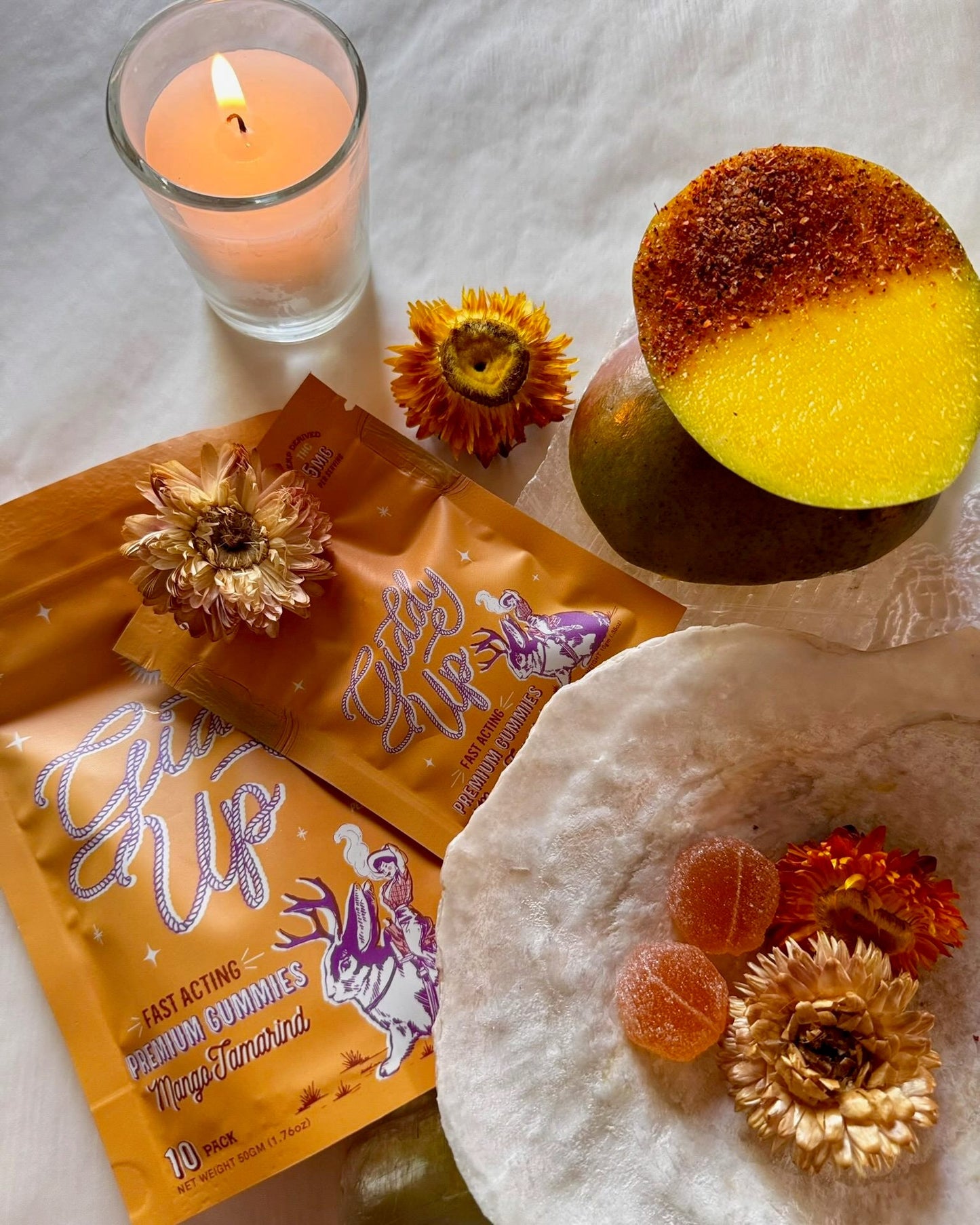 Candle, packets of 'Giddy Up' gummies, and a mango with flowers on a textured surface.