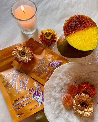 Candle, packets of 'Giddy Up' gummies, and a mango with flowers on a textured surface.